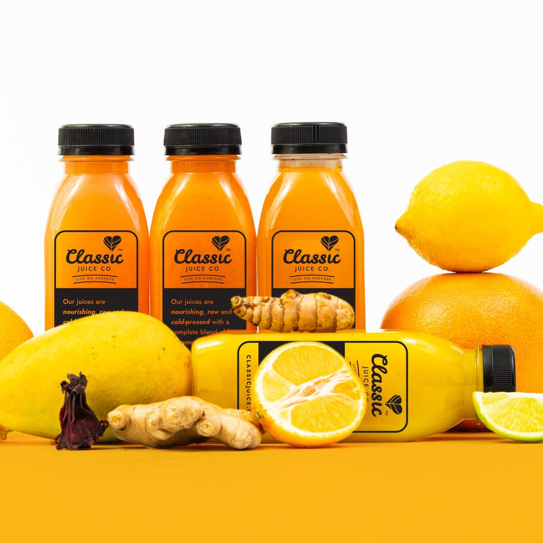 Four bottles of Classic cleansing juices. Three orange coloured juices and one yellow juice, lying in front of the other juices. Fresh fruit and veg intermixed, including a mango, lemon and ginger.