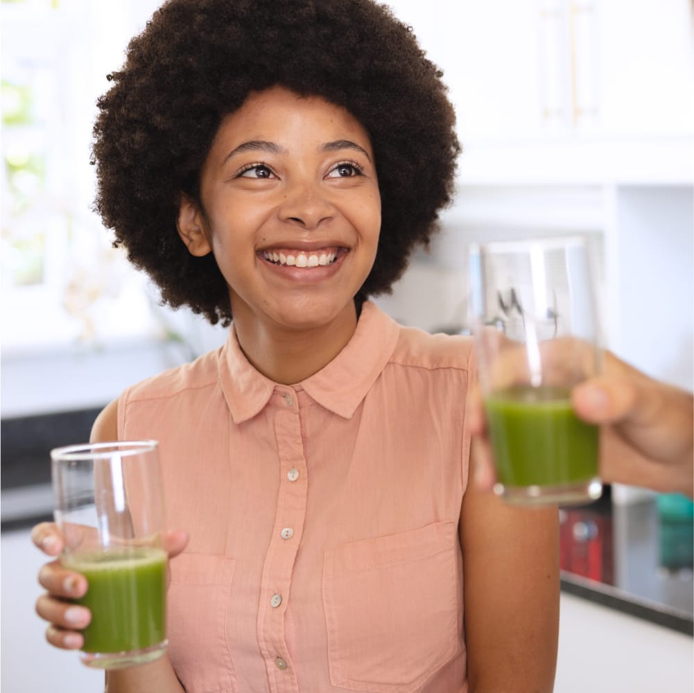 A smiling woman holding a glass of Classic cleansing green juice. The woman is wearing a peach coloured top. Another person's hand is also holding a glass of Classic cleansing green juice. The background is a kitchen setting.
