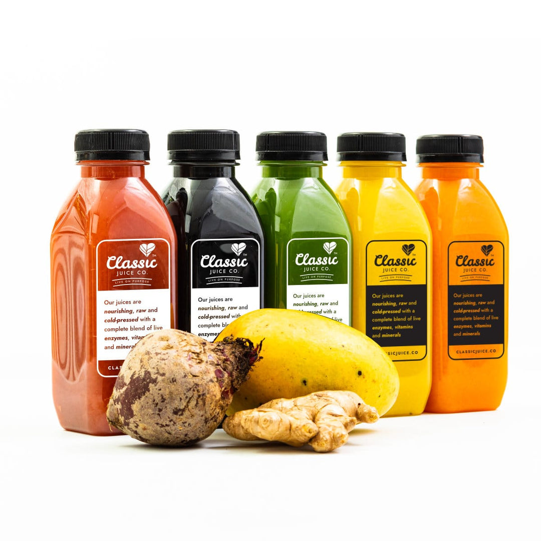 Five bottles of Classic fresh cleansing juices, all different colours: red, black, green. yellow and orange. In front of the juices is a mango, beetroot and ginger. The juices and fruit are sent against a white background.