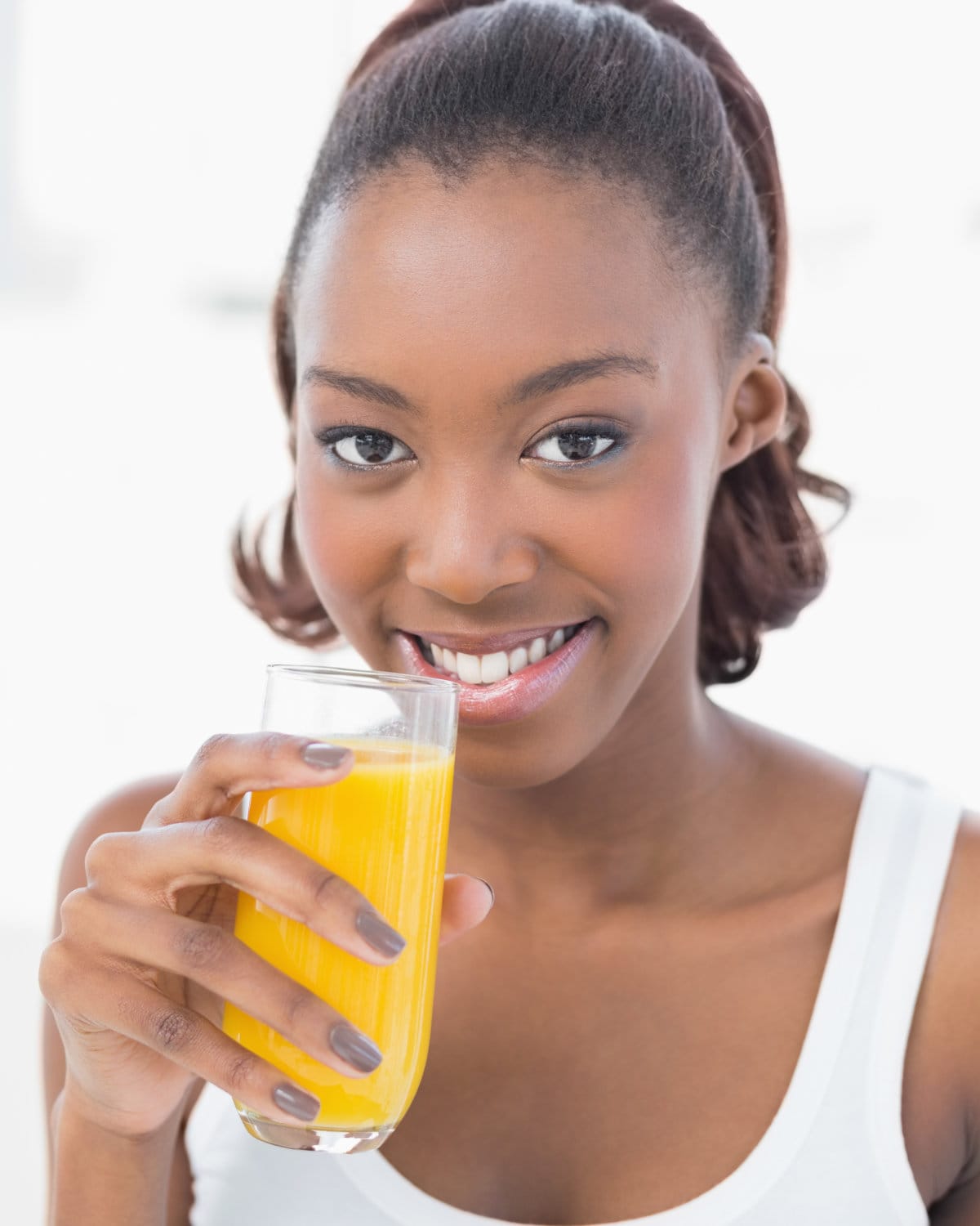 A smiling woman in a white tank top holding a glass of Classic cleansing orange juice. The background is soft focus white. 