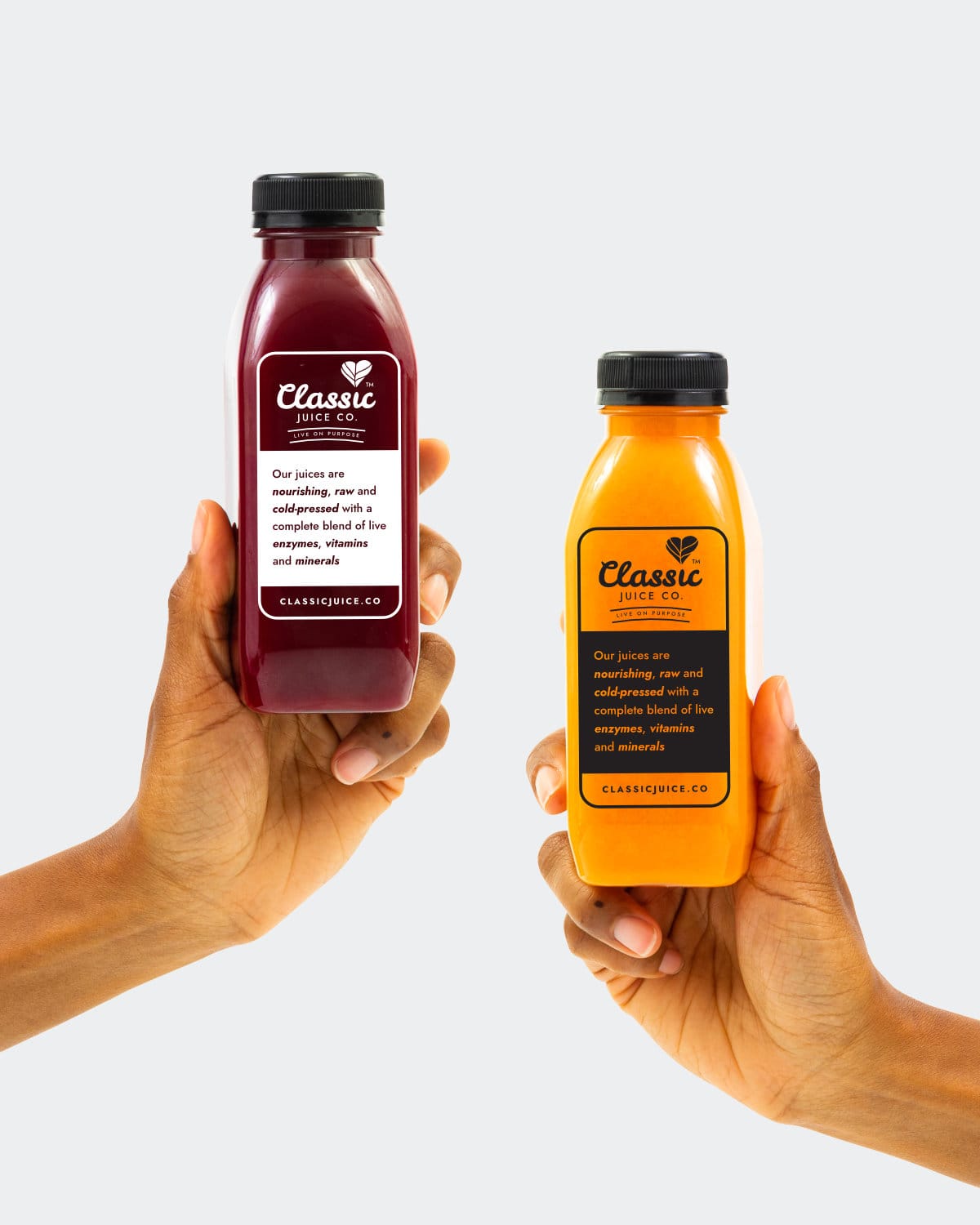 Two hands, each holding a different Classic cleansing juice. One orange and one beetroot colour. The hands and juices are set against a pale grey background.