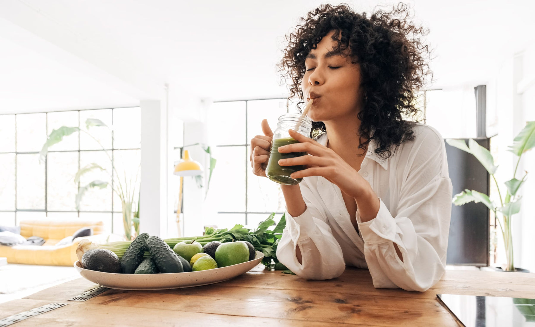 A woman enjoying a green Classic cleaning juice in a glass jar using a straw. Next to the woman is a plate of green fruit and vegetables, including an avocado, apple and lime. The woman is wearing a white shirt and sitting in a home setting.